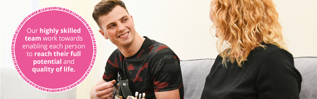 Image of a person we support playing his guitar with his support worker. He is smiling and looking at her. There is a pink roundel graphic with text that reads 'Our highly skilled team work towards enabling each person to reach their full potential and quality of life'.