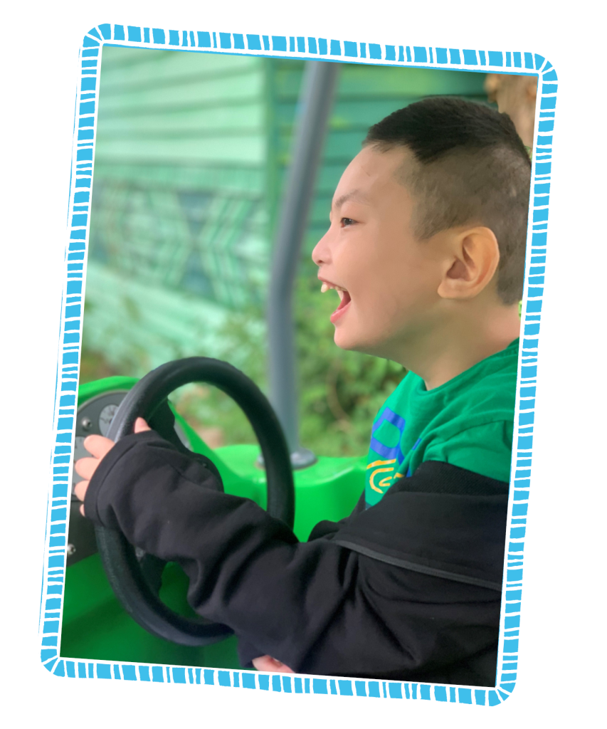 Image of Hao Lin facing the left and smiling as he drives a green toy car. He is in a green top and has a black jacket on.